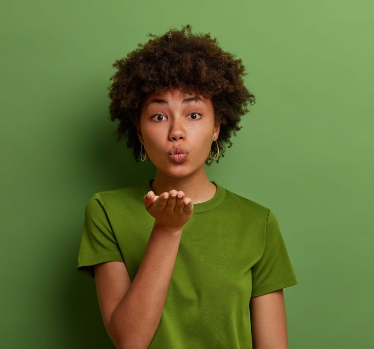 Portrait of romantic curly girlfriend sends air kiss from open palm, looks with romance, expresses affection, says be my love, kisses with pout lips, wears green t shirt. Body language concept